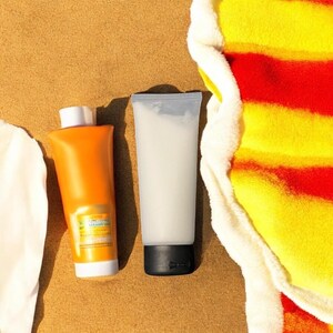 May include: Two tubes of sunscreen and a beach towel on a sandy surface. One tube is orange with a white cap, the other is translucent with a black cap. The towel has red, yellow, and white stripes.