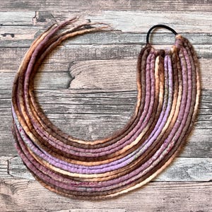 Könnte beinhalten: Handgefertigte Dreadlocks in Braun-, Lila- und Beigetönen, kreisförmig angeordnet. Die Dreadlocks sind an einem schwarzen Gummiband befestigt. Die Textur ist weich und faserig, mit Farb- und Dickenvariationen.