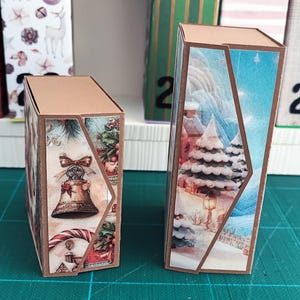 Peut inclure: Deux petites boîtes rectangulaires avec des bords et des dessus bruns. Les boîtes présentent des panneaux décoratifs avec des illustrations sur le thème de Noël. L'une montre une cloche et un sucre d'orge, l'autre une scène hivernale enneigée.