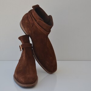 Pode incluir: Um par de botas de tornozelo de camur&ccedil;a marrom com fecho de fivela. As botas t&ecirc;m bico fino e salto baixo.