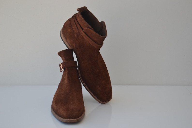Pode incluir: Um par de botas de tornozelo de camur&ccedil;a marrom com fecho de fivela. As botas t&ecirc;m bico fino e salto baixo.