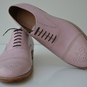 May include: A pair of pink leather oxford shoes with brown laces. The shoes have a brogue design with a heart-shaped pattern on the toe.