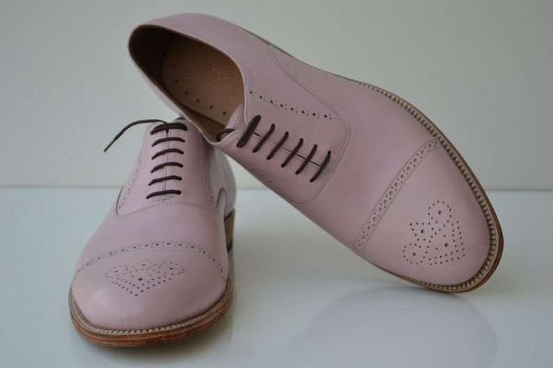 May include: A pair of pink leather oxford shoes with brown laces. The shoes have a brogue design with a heart-shaped pattern on the toe.
