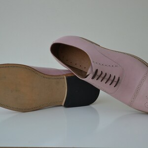 May include: A pair of pink leather oxford shoes with a brogue design and a brown leather sole.