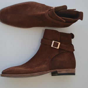 Pode incluir: Um par de botas de tornozelo de camur&ccedil;a marrom com uma fivela. As botas t&ecirc;m uma ponta pontiaguda e um salto baixo.
