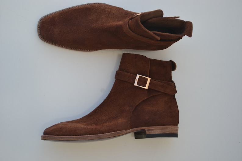Pode incluir: Um par de botas de tornozelo de camur&ccedil;a marrom com uma fivela. As botas t&ecirc;m uma ponta pontiaguda e um salto baixo.