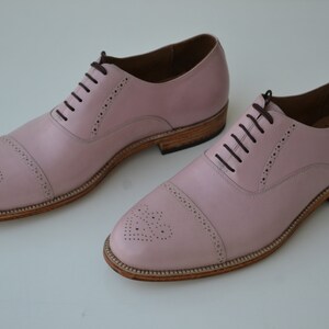 May include: A pair of pink leather lace-up shoes with a brogue design and a brown leather sole.