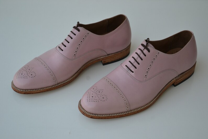 May include: A pair of pink leather lace-up shoes with a brogue design and a brown leather sole.