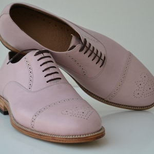 May include: A pair of pink leather lace-up shoes with a brogue design. The shoes have a rounded toe and a brown leather sole.