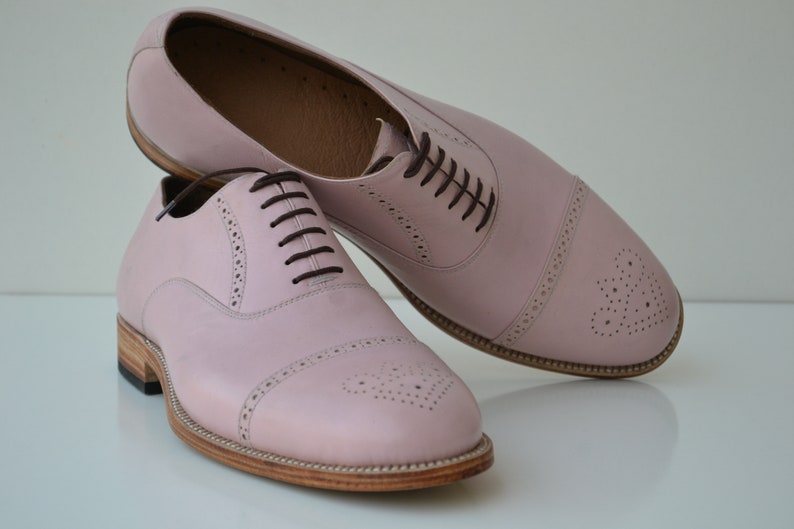 May include: A pair of pink leather lace-up shoes with a brogue design. The shoes have a rounded toe and a brown leather sole.