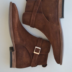 Pode incluir: Um par de botas de tornozelo de camur&ccedil;a marrom com uma fivela. As botas t&ecirc;m bico fino e salto baixo.