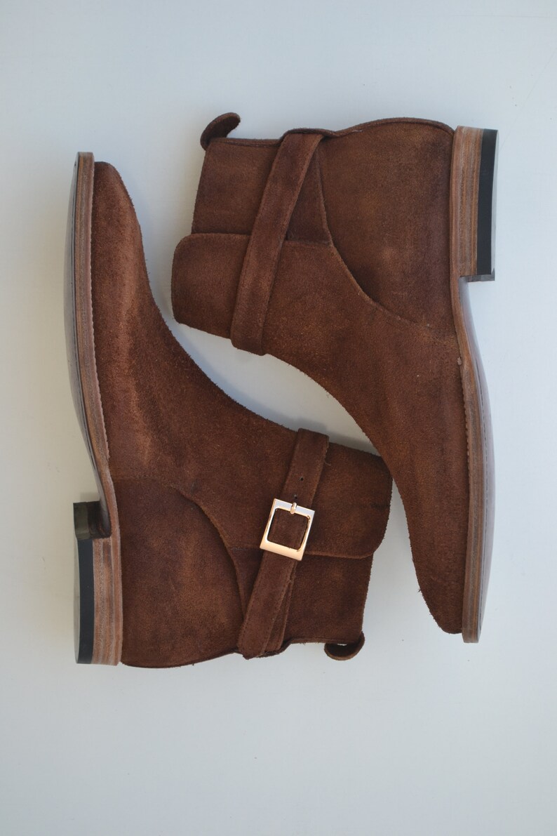 Pode incluir: Um par de botas de tornozelo de camur&ccedil;a marrom com uma fivela. As botas t&ecirc;m bico fino e salto baixo.