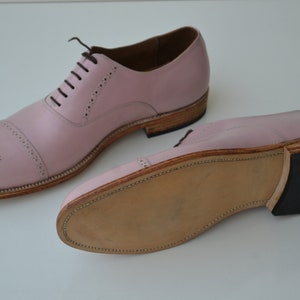 May include: A pair of pink leather lace-up shoes with a brogue design. The shoes have a brown leather sole and a black rubber heel.