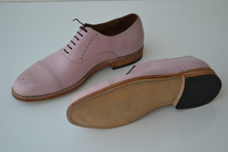 May include: A pair of pink leather lace-up shoes with a brogue design. The shoes have a brown leather sole and a black rubber heel.