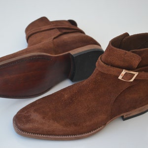Pode incluir: Um par de botas de tornozelo de camur&ccedil;a marrom com uma fivela. As botas t&ecirc;m bico fino e salto baixo.