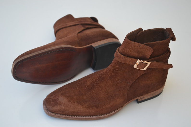 Pode incluir: Um par de botas de tornozelo de camur&ccedil;a marrom com uma fivela. As botas t&ecirc;m bico fino e salto baixo.