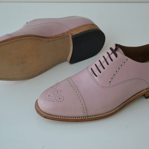May include: A pair of pink leather lace-up shoes with a brogue design. The shoes have a brown leather sole and a black rubber heel.