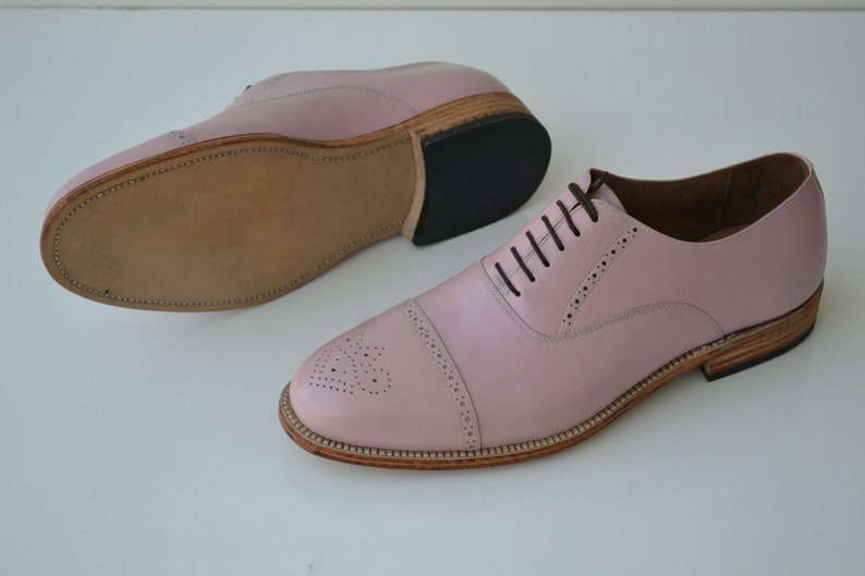 May include: A pair of pink leather lace-up shoes with a brogue design. The shoes have a brown leather sole and a black rubber heel.