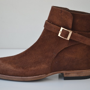 Pode incluir: Botas de tornozelo de camur&ccedil;a marrom com uma fivela dourada.