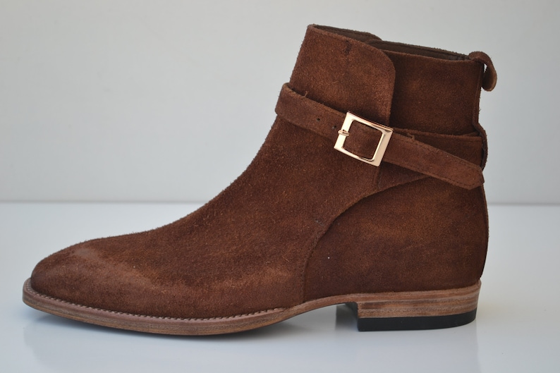 Pode incluir: Botas de tornozelo de camur&ccedil;a marrom com uma fivela dourada.