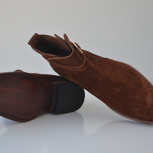 Pode incluir: Um par de botas de tornozelo de camur&ccedil;a marrom com fecho de fivela. As botas t&ecirc;m bico fino e salto baixo.