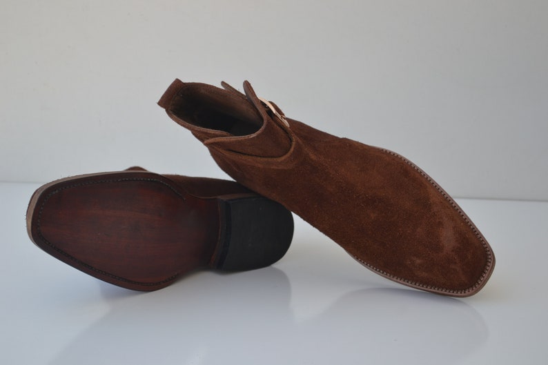 Pode incluir: Um par de botas de tornozelo de camur&ccedil;a marrom com fecho de fivela. As botas t&ecirc;m bico fino e salto baixo.