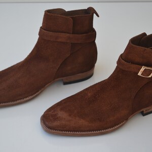 Pode incluir: Um par de botas de tornozelo de camur&ccedil;a marrom com uma fivela. As botas t&ecirc;m uma ponta pontiaguda e um salto baixo.