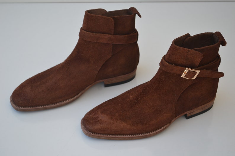 Pode incluir: Um par de botas de tornozelo de camur&ccedil;a marrom com uma fivela. As botas t&ecirc;m uma ponta pontiaguda e um salto baixo.