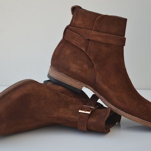 Pode incluir: Botas de tornozelo de camur&ccedil;a marrom com uma fivela e bico fino.