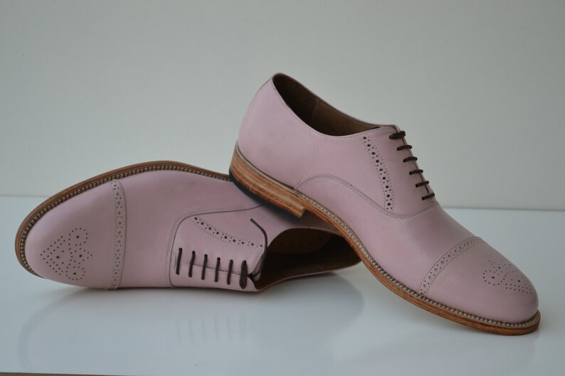May include: A pair of pink leather lace-up shoes with brogue detailing. The shoes have a rounded toe and a low heel.