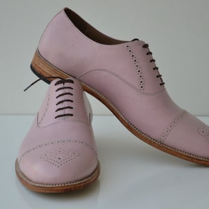 May include: A pair of pink leather lace-up shoes with a brogue design. The shoes have a brown leather sole and laces.