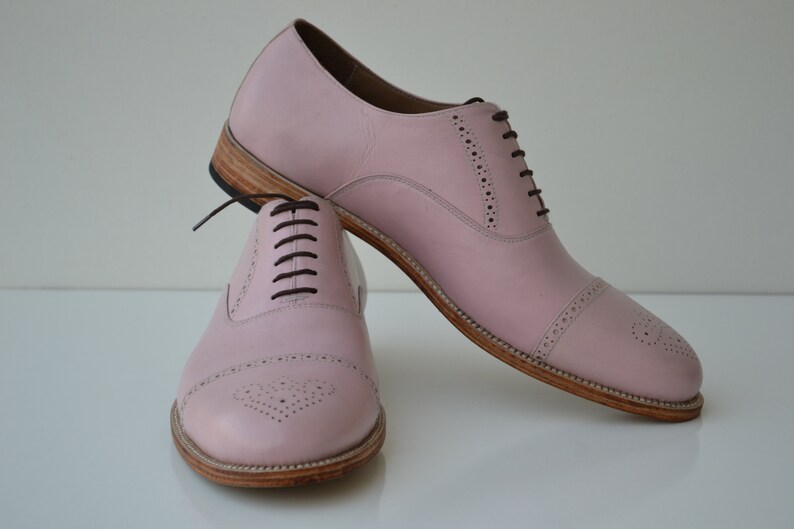 May include: A pair of pink leather lace-up shoes with a brogue design. The shoes have a brown leather sole and laces.