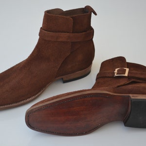 Pode incluir: Botas de tornozelo de camur&ccedil;a marrom com uma fivela e uma sola de couro.