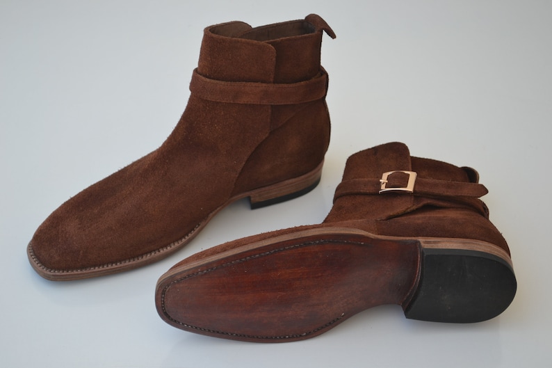Pode incluir: Botas de tornozelo de camur&ccedil;a marrom com uma fivela e uma sola de couro.