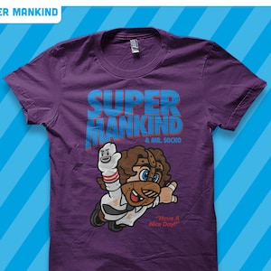Könnte beinhalten: Lilafarbenes T-Shirt mit einer Comicfigur, die eine Maske und ein weißes Hemd trägt. Der Text "SUPER MANKIND & MR. SOCKO" ist auf dem T-Shirt aufgedruckt. Die Figur hält einen Handschuh mit einem Smiley-Gesicht. Der Text "Have A Nice Day!" ist unter der Figur aufgedruckt.