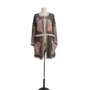May include: A long, open-front coat in shades of gray, brown, and pink, displayed on a mannequin. The coat features patchwork details, floral appliques, and ruffled trim. The sleeves are long, and the coat has two pockets. The overall style is bohemian.