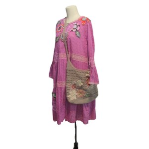 May include: A pink dress with floral embroidery and a crochet shoulder bag. The dress has a loose fit with bell sleeves and lace trim. The bag is beige with floral embellishments.