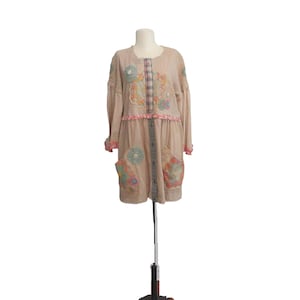 May include: A beige, long-sleeved dress with floral and patchwork details. The dress features colorful embroidered patches, decorative buttons, and a ruffled hem. The dress is displayed on a mannequin.