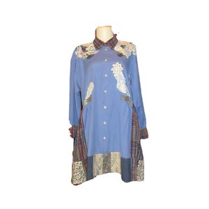 May include: A long-sleeved blue shirt with patchwork details. The shirt features a plaid collar and cuffs, with floral and patterned fabric accents. White floral appliqués are sewn onto the front of the shirt. The shirt is displayed on a mannequin.