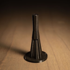 May include: A black, decorative bottle stopper with a unique, geometric design. The stopper has a flared base, a ridged, conical body, and a slender, faceted top. The object is made of a dark material and sits on a wooden surface.