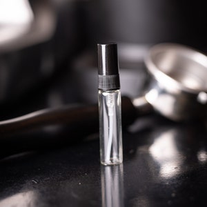 May include: A small clear glass bottle with a black spray nozzle, sitting on a black surface. The bottle is filled with a clear liquid.