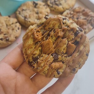 May include: A close-up of a chocolate chip cookie with caramel drizzle and chocolate chips. The cookie is being held in a hand. The cookie is on a white surface. The text "Cakespellian" is visible in the background.
