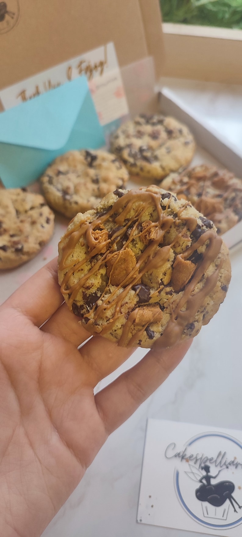 May include: A close-up of a chocolate chip cookie with caramel drizzle and chocolate chips. The cookie is being held in a hand. The cookie is on a white surface. The text "Cakespellian" is visible in the background.