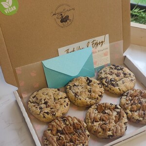 May include: A white box with six chocolate chip cookies. The cookies are arranged in a circle. The box is lined with white paper and has a pink and white pattern. The box is open and the cookies are visible. The box is labeled "Vegan" and "Cakespellintimes".