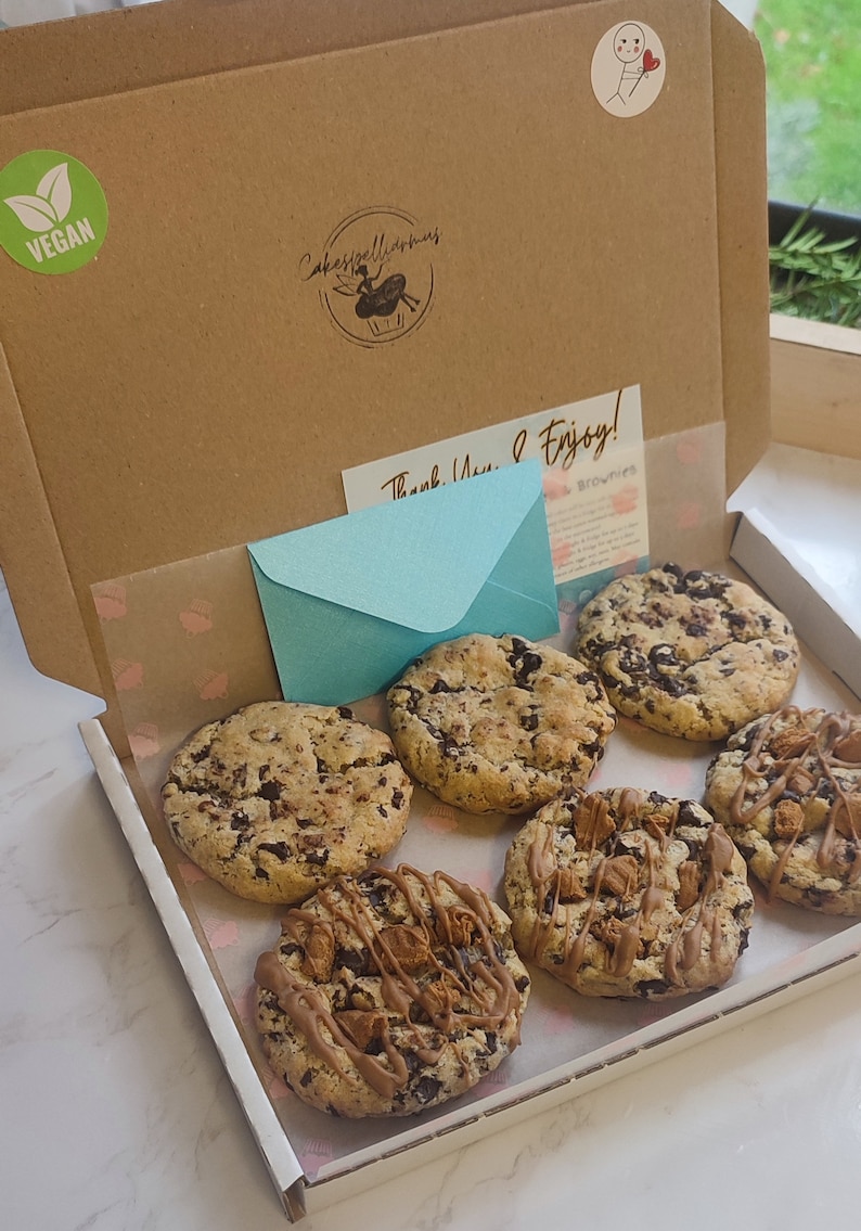 May include: A white box with six chocolate chip cookies. The cookies are arranged in a circle. The box is lined with white paper and has a pink and white pattern. The box is open and the cookies are visible. The box is labeled "Vegan" and "Cakespellintimes".