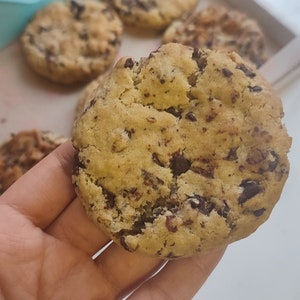 May include: Close-up of a large, golden brown chocolate chip cookie with dark chocolate chips. The cookie is being held in a hand. The cookie is in focus, and the background is blurred.