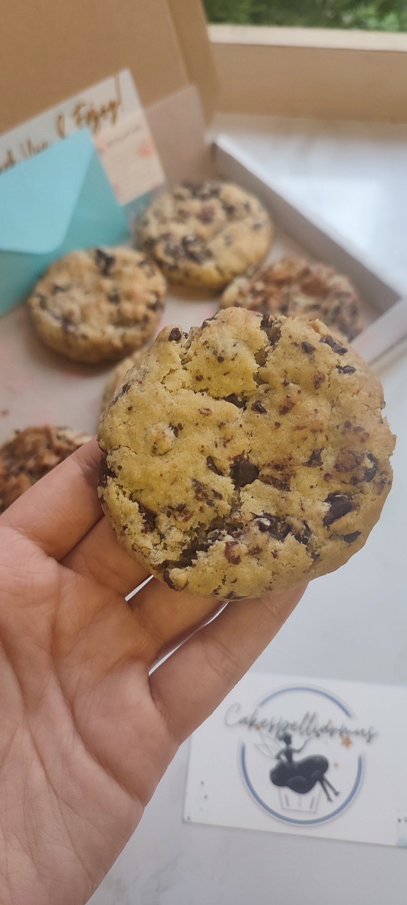 May include: Close-up of a large, golden brown chocolate chip cookie with dark chocolate chips. The cookie is being held in a hand. The cookie is in focus, and the background is blurred.