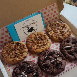 May include: A box of six vegan cookies. The cookies are arranged in a circle on a pink and white heart-shaped paper. The cookies are a variety of flavors, including chocolate chip, peanut butter, and oatmeal raisin. The box is labeled "Cakespelliarmus" and "Vegan".