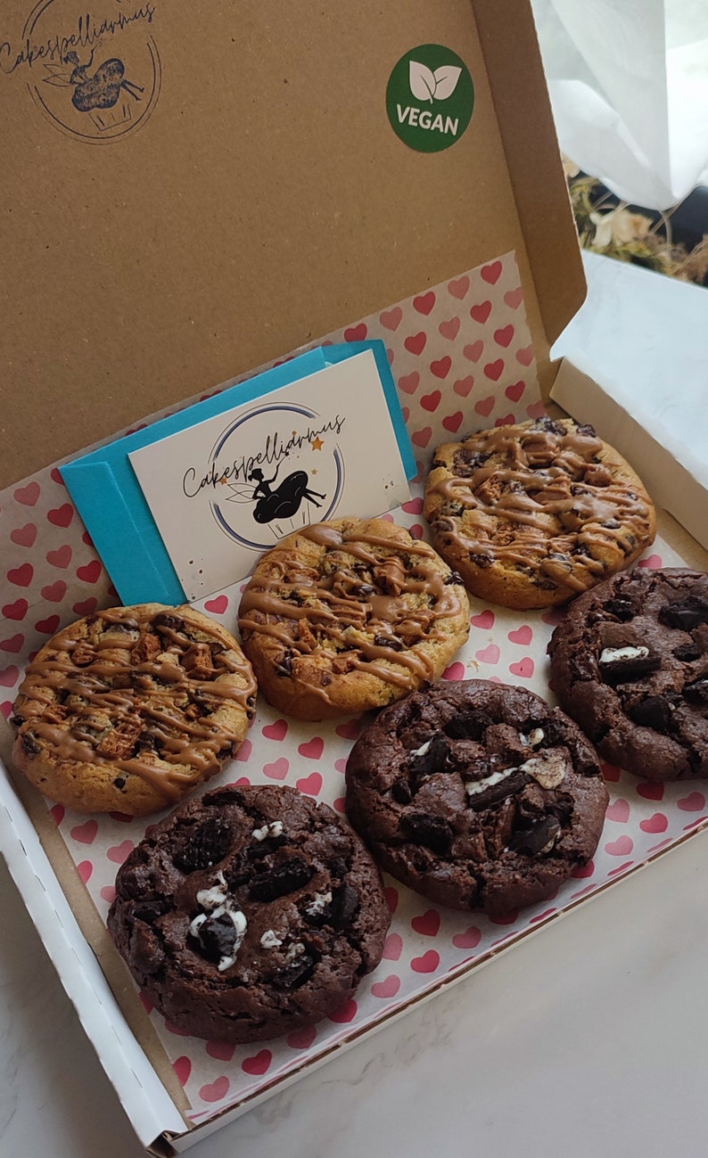 May include: A box of six vegan cookies. The cookies are arranged in a circle on a pink and white heart-shaped paper. The cookies are a variety of flavors, including chocolate chip, peanut butter, and oatmeal raisin. The box is labeled "Cakespelliarmus" and "Vegan".