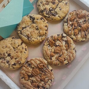 May include: A box of six assorted cookies. The cookies are a mix of chocolate chip and chocolate chip with caramel and chocolate drizzle. The box is lined with white paper with a pink cupcake pattern. The box has a handwritten note that says "Thank You & Enjoy!"